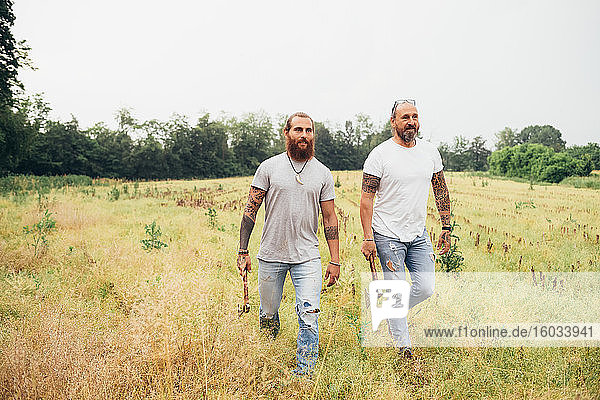 Two bearded tattooed men with long brunette hair walking across a meadow  carrying axes.