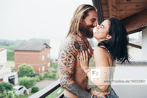Bärtiger tätowierter Mann mit langen brünetten Haaren und Frau mit langen braunen Haaren stehen auf einem Balkon  umarmen sich und küssen sich.