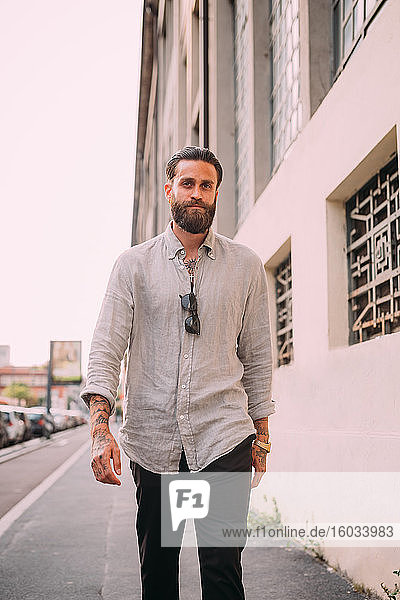 Portrait of bearded young man with brown hair  with tattoos on arms  wearing grey shirt  walking down street.