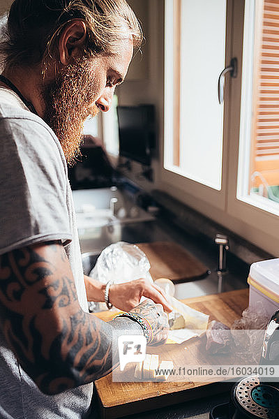Bärtiger tätowierter Mann mit langen brünetten Haaren  der in einer Küche steht und Essen zubereitet.