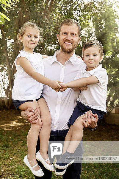 Familienporträt  Mann steht im Freien  hält Junge und Mädchen in der Hand und lächelt in die Kamera.