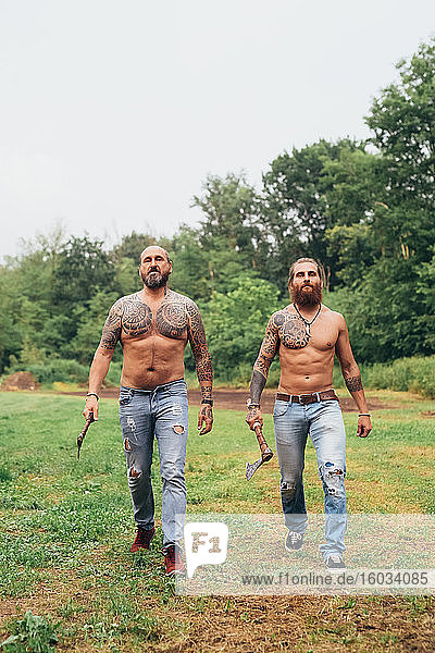 Two bearded tattooed men with long brunette hair  shirtless and wearing jeans  walking across a meadow  carrying axes.