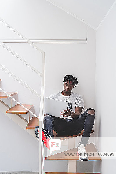 Junger Mann mit kurzen Dreadlocks  der auf einer Treppe sitzt und am Laptop tippt.