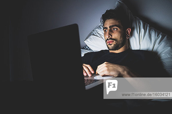 Bearded young man lying in bed at night  looking at laptop.