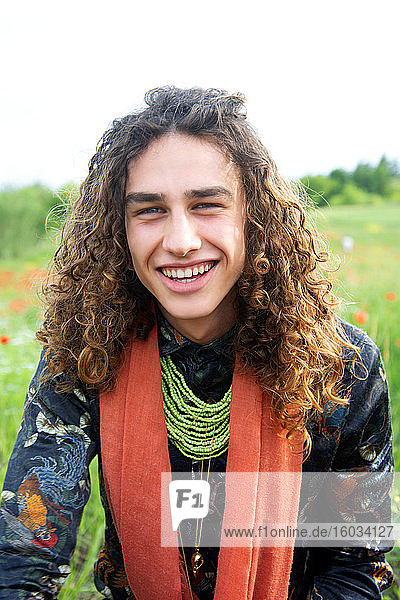 Portrait of young man with long brown curly hair in a poppy meadow  smiling at camera.