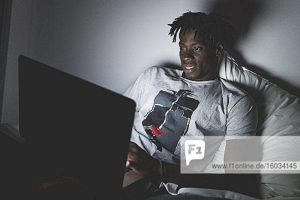 Young man with short dreadlocks lying in bed at night  looking at laptop.