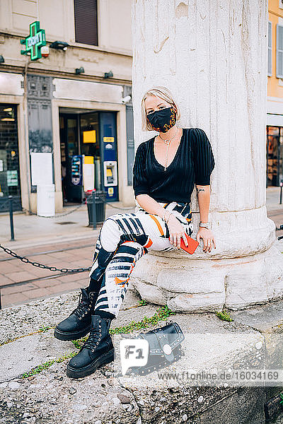 Junge Frau  die während des Coronavirus eine Gesichtsmaske trägt  sitzt im Freien und schaut in die Kamera.