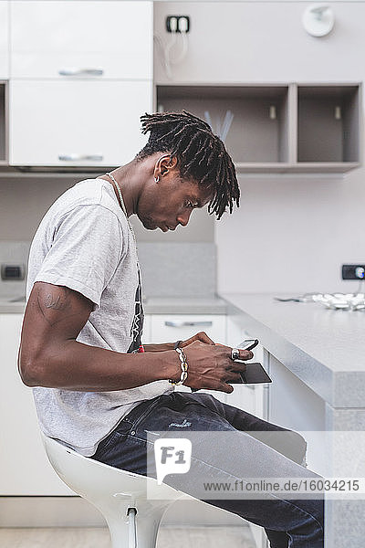 Young man with short dreadlocks sitting on bar stool in kitchen  using mobile phone.