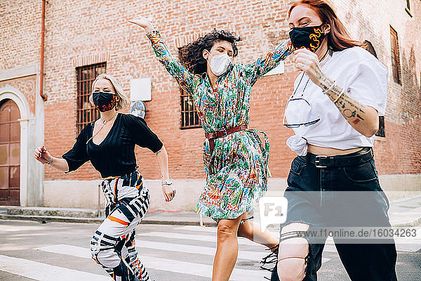 Drei junge Frauen mit Gesichtsmasken während des Corona-Virus  die in einer Straße über einen Fußgängerüberweg rannten.