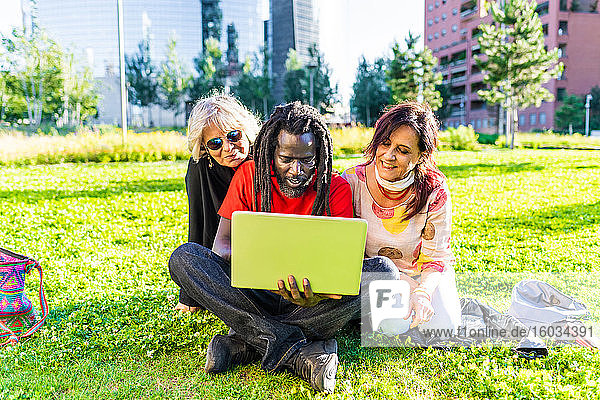 Schwarzer Mann mit Dreadlocks und zwei kaukasische Frauen sitzen auf dem Rasen und schauen auf einen Laptop.