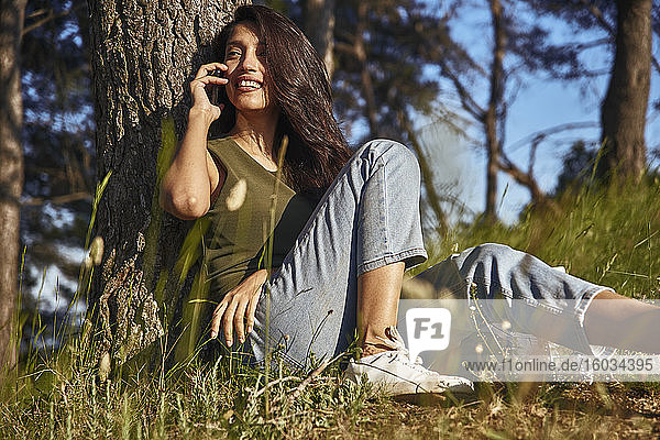 Porträt einer jungen Frau mit langen braunen Haaren  die unter einem Baum im Wald sitzt und mit dem Handy telefoniert.
