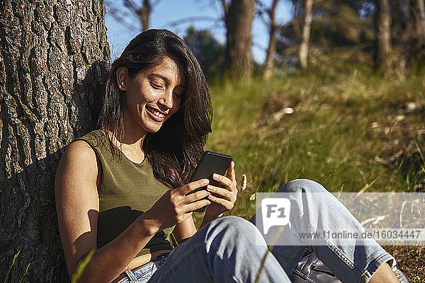 Porträt einer jungen Frau mit langen braunen Haaren  die unter einem Baum in einem Wald sitzt und ihr Handy überprüft.