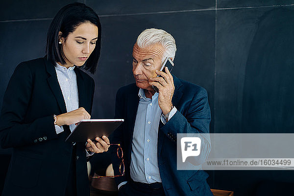 Geschäftsmann und -frau betrachten digitales Tablet und telefonieren mit Smartphone im Sitzungssaal