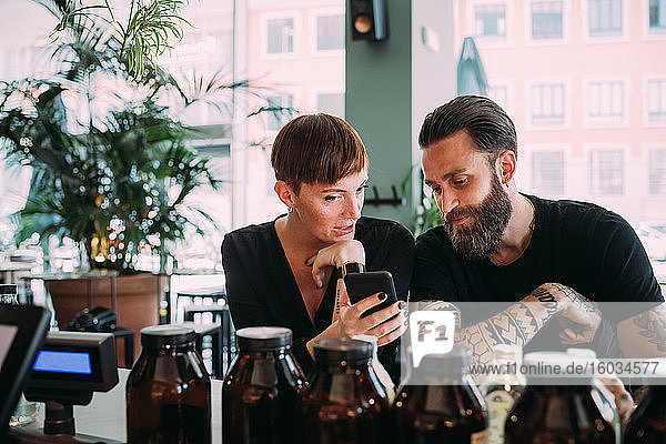 Bearded young man with brown hair and tattoos and young woman with short hair sitting in a bar  looking at mobile phone.