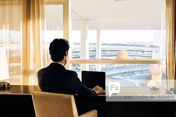 Businessman using laptop and looking out of hotel bedroom window