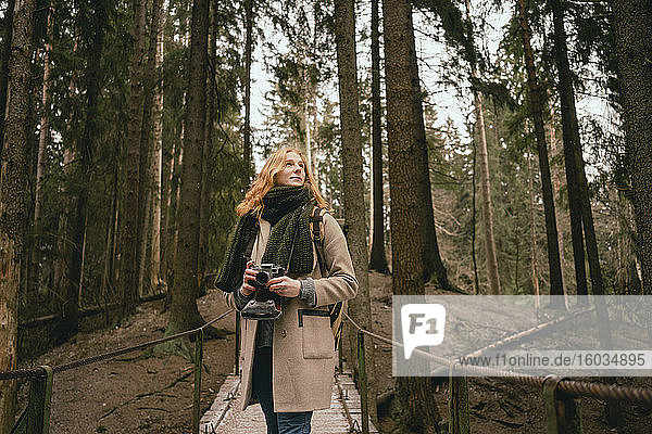 Gelassene rothaarige Frau mit Kamera auf Waldsteg