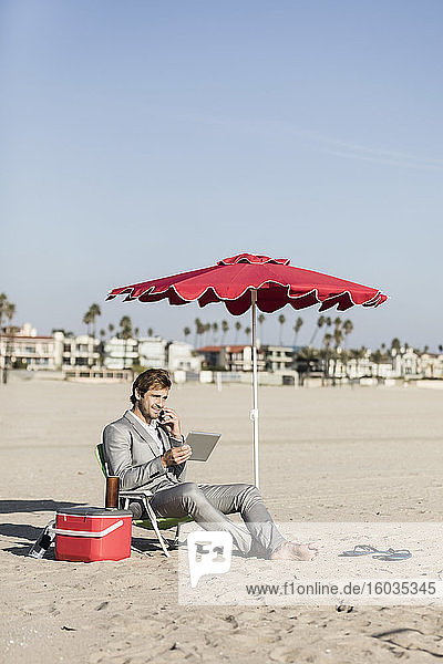 Barfüßiger Geschäftsmann mit Smartphone und digitalem Tablet arbeitet am Strand  Los Angeles  Kalifornien