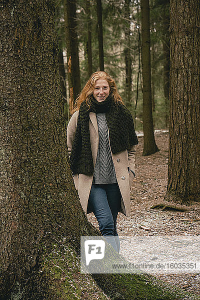 Porträt einer selbstbewussten rothaarigen Frau in Mantel und Schal zwischen Bäumen im Wald stehend