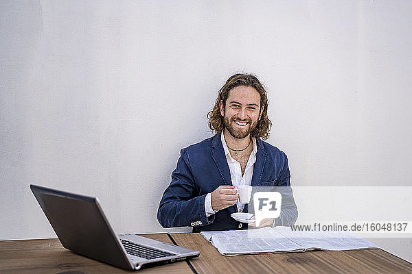 Glücklicher männlicher Jungunternehmer sitzt mit Espresso und Zeitung an der weißen Wand eines Cafés