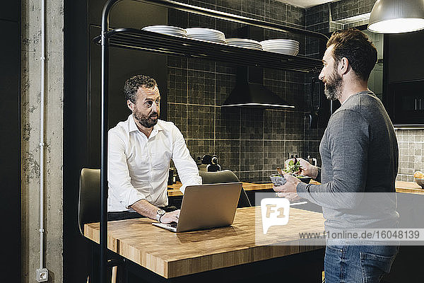 Mature friends working on laptop in kitchen  one eating salad