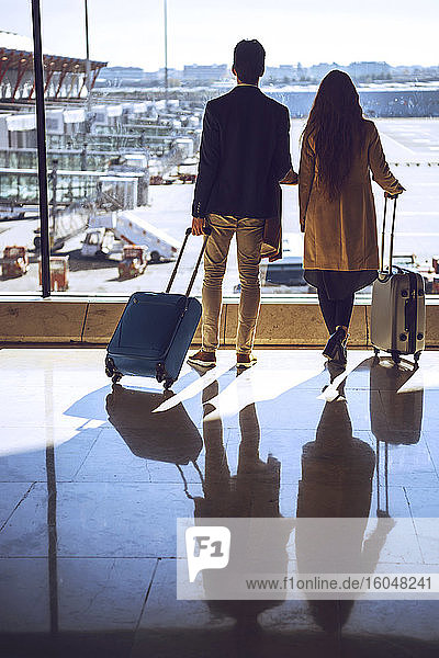 Business couple holding luggage while looking through window at airport departure area