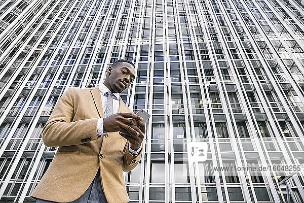 Junger Geschäftsmann vor einem modernen Bürogebäude in der Stadt  der sein Smartphone benutzt