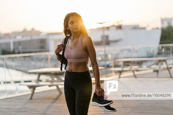 Nachdenkliche Sportlerin mit Schuhen in der Hand  während sie bei Sonnenuntergang am Pier steht