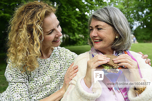Lächelnde blonde Frau betrachtet ältere Mutter beim Essen eines Sandwiches im Park