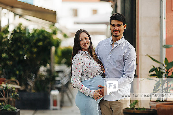 Glückliches schwangeres Paar mit Händen auf dem Bauch steht in der Stadt während des sonnigen Tages
