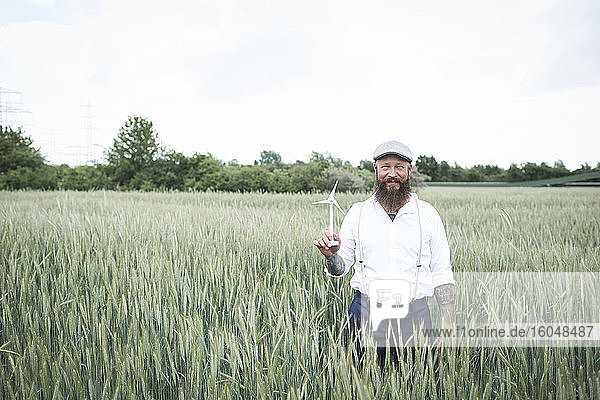 Bärtiger Mann  der eine kleine Windmühle hält  während er inmitten eines Kornfelds vor dem Himmel steht