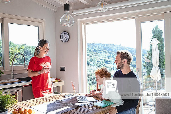 Lächelnde Eltern  die sich gegenseitig ansehen  während ihr Sohn in der Küche einen Laptop benutzt