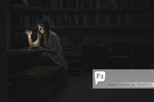 Young woman sitting in office  reading book at night with a torch