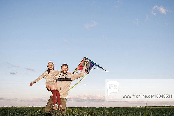 Mann  der einen Drachen hält und seine Tochter glücklich trägt  während er in einer grünen Landschaft steht