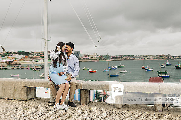 Romantisches schwangeres Paar sitzt auf einer Stützmauer am Hafen  Cascais  Portugal