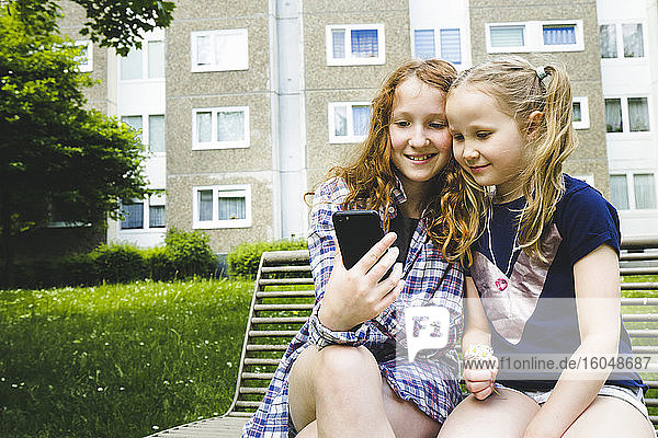 Lächelnde Geschwister teilen sich ein Smartphone  während sie auf einer Bank im Park sitzen