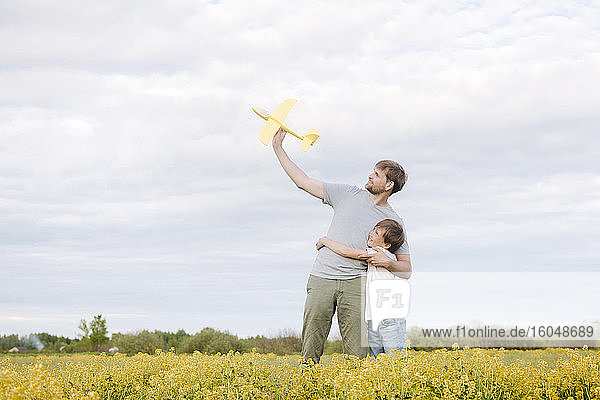 Lächelnder Junge  der seinen Vater umarmt und ein Spielzeugflugzeug auf einem Rapsfeld gegen den Himmel hält
