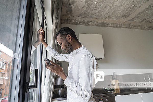 Nachdenklicher Mann an der Terrassentür seines Hauses mit Mobiltelefon