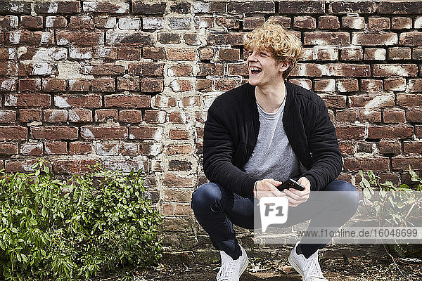 Porträt eines lachenden jungen Mannes mit Smartphone  der vor einer Backsteinmauer kaut