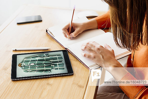 Weibliche Modedesignerin arbeitet zu Hause am Schreibtisch sitzend mit Tablet und macht sich Notizen
