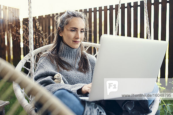 Schöne junge Frau  die einen Laptop benutzt und auf einer Schaukel im Garten sitzt