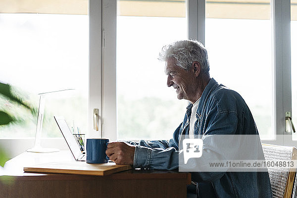 Glücklicher älterer Mann  der eine Kaffeetasse hält  während er einen Laptop zu Hause benutzt