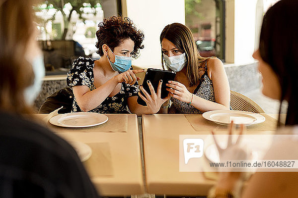 Frauen tragen Masken  während sie mit Freunden an einem Tisch im Restaurant sitzen und ein digitales Tablet benutzen
