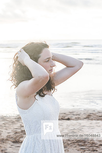 Nachdenkliche junge Frau in weißem Kleid mit Händen im Haar am Strand stehend