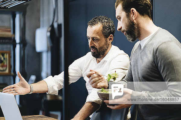 Mature friends working on laptop in kitchen  one eating salad