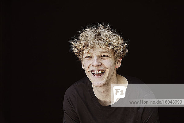 Porträt eines lachenden jungen Mannes mit lockigem blondem Haar vor schwarzem Hintergrund