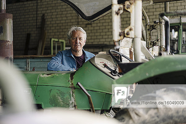 Porträt eines älteren Mannes auf einem Bauernhof mit Traktor in einer Scheune