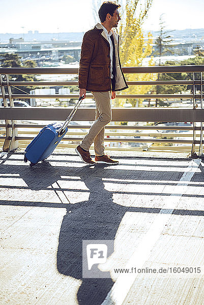 Geschäftsmann beim Ziehen des Gepäcks auf einem erhöhten Gehweg am Flughafen an einem sonnigen Tag