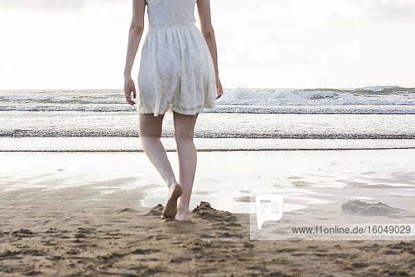 Junge Frau in weißem Kleid läuft barfuß am Strand gegen den klaren Himmel