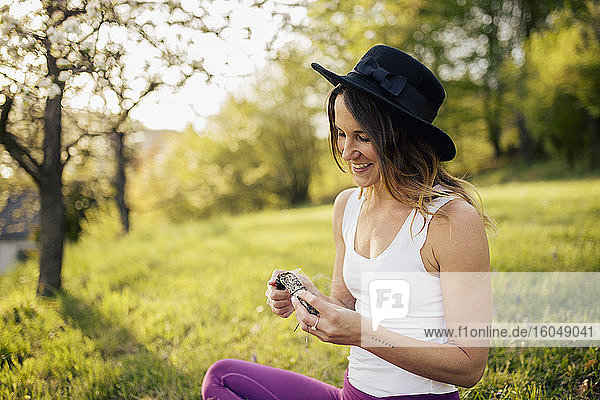 Lächelnde Frau mit Hut  die Weihrauch verbrennt  während sie auf einer Wiese im Park sitzt