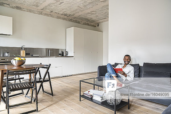Lachender Mann mit Buch auf Couch in moderner Wohnung sitzend